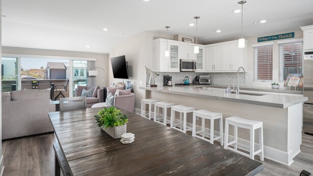 Una cocina moderna con armarios blancos, una isla central con taburetes de bar, una mesa de comedor con un centro de mesa de plantas y un televisor de pantalla plana montado en la pared.