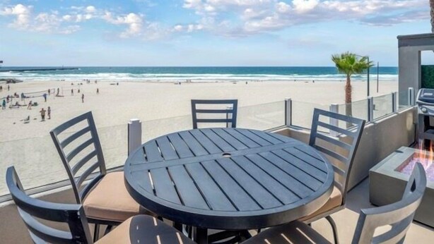 A beachside patio with a round table and chairs, overlooking a sandy beach and the ocean.