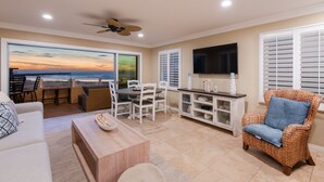 A spacious living area with a view of the ocean, a dining table with chairs, a sofa, a wicker armchair, and a flat-screen TV mounted on the wall.