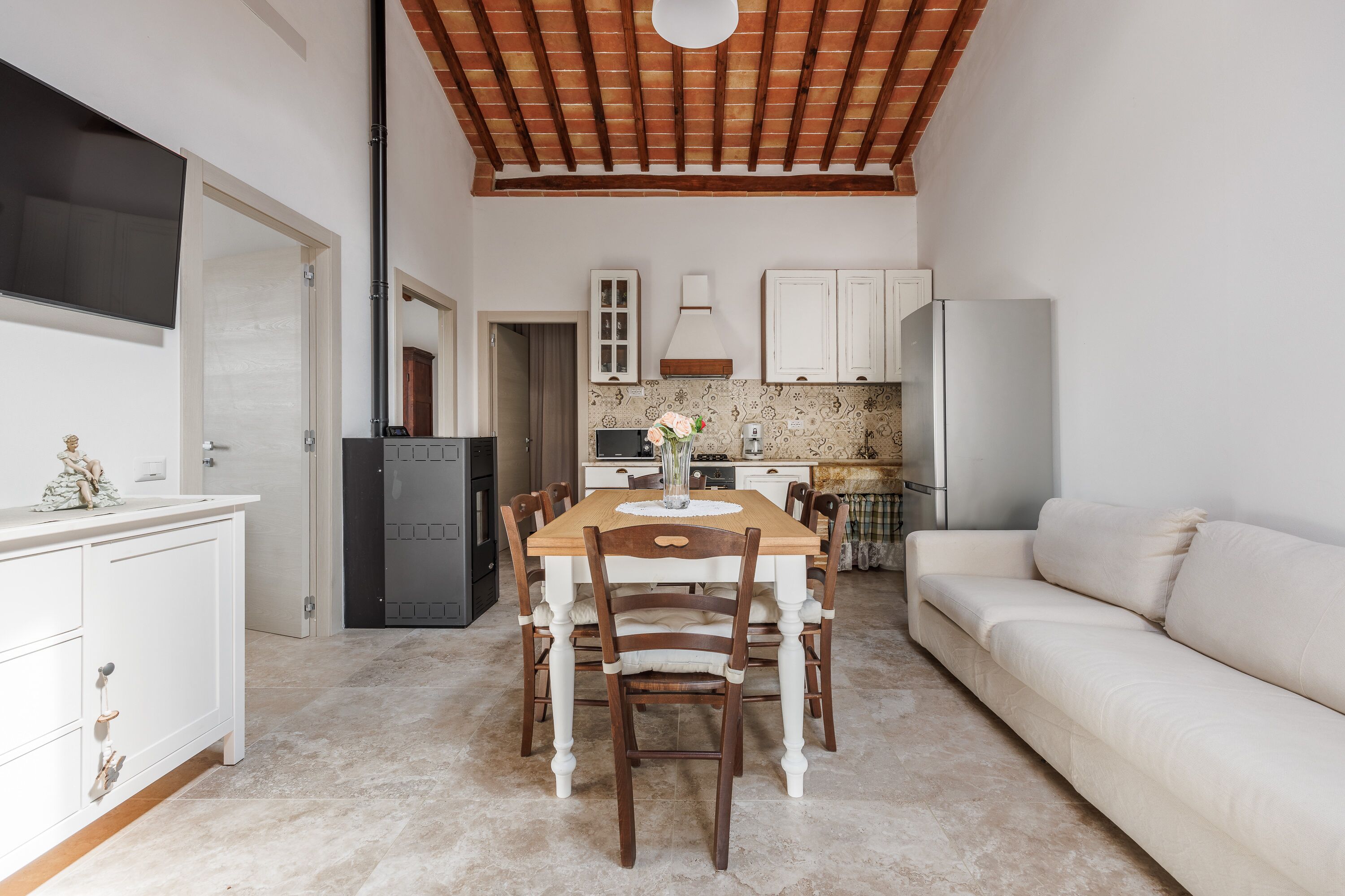 Een compact woongedeelte met een eettafel omringd door stoelen, een bank, een keuken met kasten en apparatuur, en een tv aan de wand.