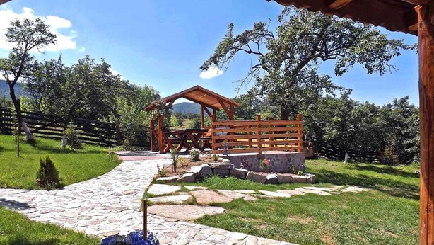 Un sendero de piedra lleva a un porche de madera con una mesa de picnic, rodeado de abundante vegetación y un muro de piedra.