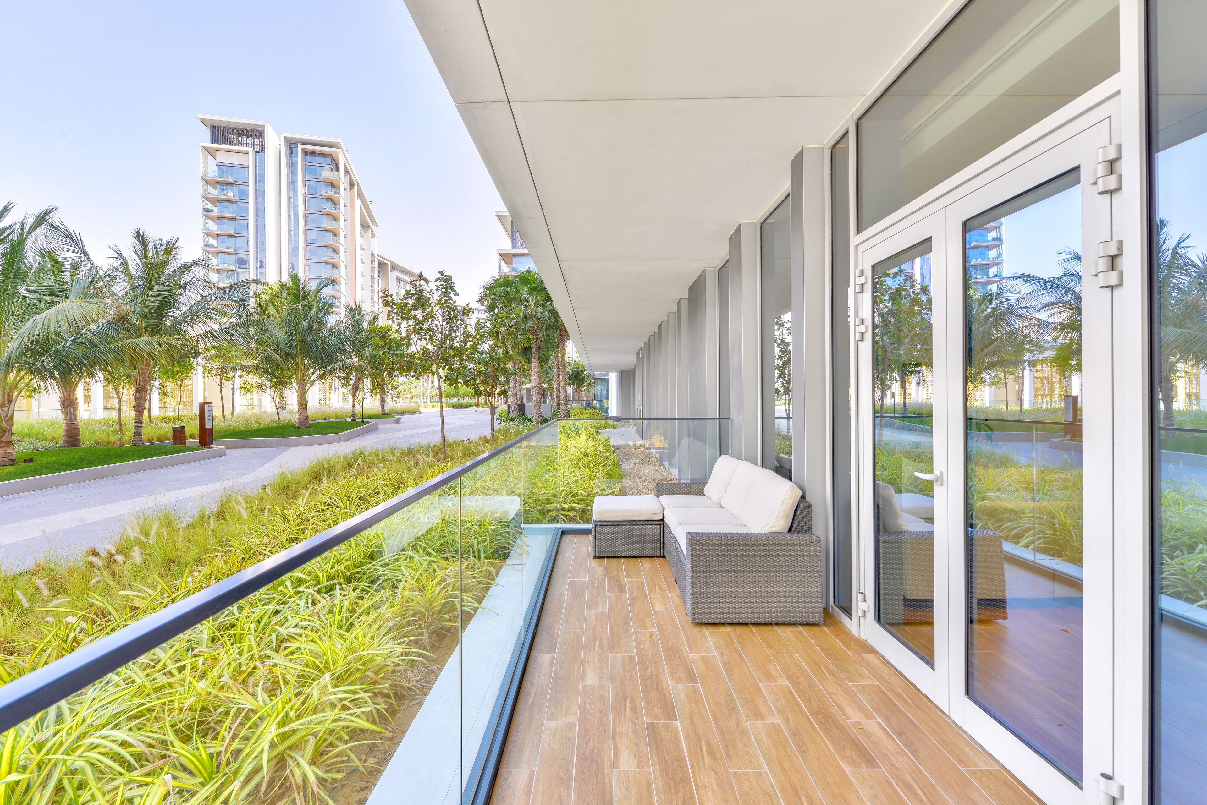 A modern balcony with a glass railing, a wicker sofa, and a view of a road with palm trees and buildings.