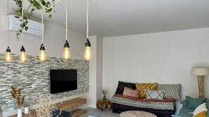 A modern living room with a stone wall, a flat-screen TV, hanging pendant lights, a sofa with patterned cushions, and a dining area with a wooden table and chairs. - Les terrasses de valerian (Brignoles)