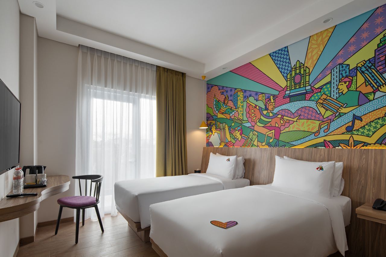 Habitación de hotel con dos camas, un escritorio, una silla y un mural colorido en la pared.