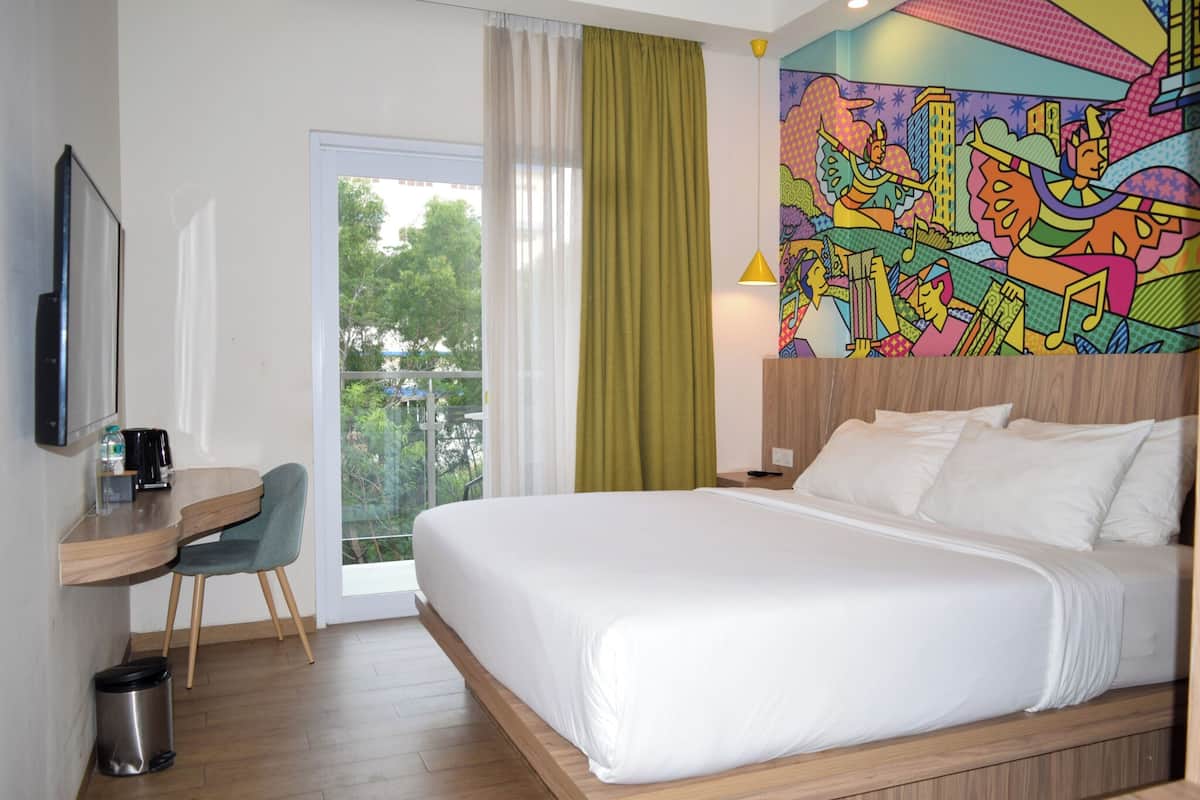 Une chambre d’hôtel équipée d’un lit, d’un bureau, d’une chaise, d’une poubelle, d’une fenêtre avec des rideaux et d’une fresque colorée sur le mur.