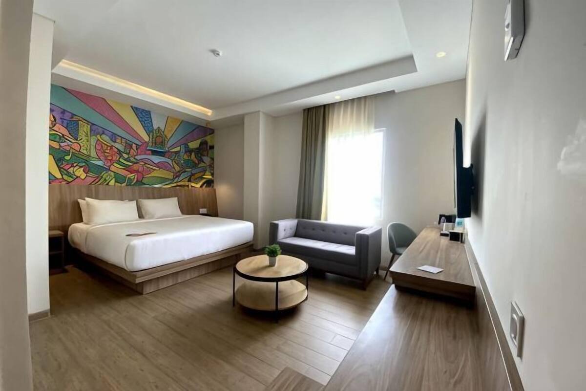Ein Hotelzimmer mit einem Bett, einem Sofa, einem Couchtisch, einem Fernseher und einem großen, farbenfrohen Wandgemälde.