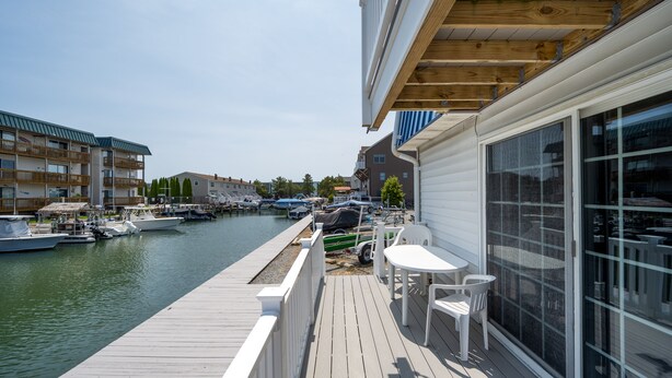 A wooden deck with a white railing, a table, and chairs, overlooking a marina with boats. - Two Bedroom Townhome On Canal! (Ocean City)