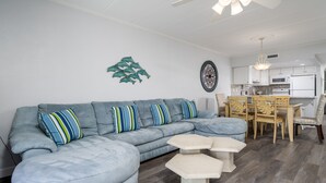A modern living room with a sectional sofa, a dining table with chairs, and a kitchen area in the background. - Oceanfront 2 bedroom in desirable North OC (Ocean City)