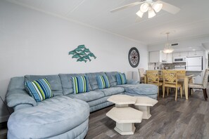 Uma sala de estar moderna com um sofá seccional, uma mesa de jantar com cadeiras e uma cozinha ao fundo.