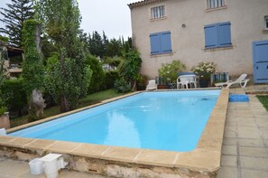 Piscina retangular com borda de pedra, rodeada por vegetação e um edifício com persianas azuis.