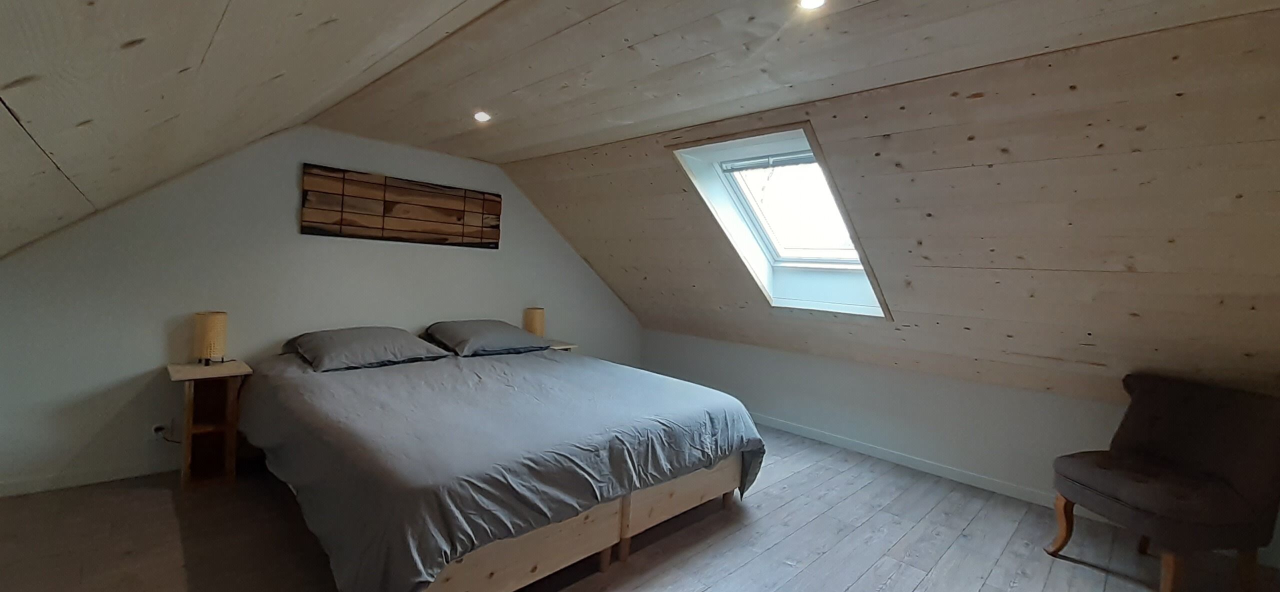 Un dormitorio con techo inclinado, tragaluz, cama con ropa de cama gris, dos mesitas de noche con lámparas y una silla.