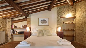 Ein Schlafzimmer mit einem großen Bett, Holzbalken, Steinwänden, einem Kamin aus Ziegeln und einem Regal mit Dekoration.