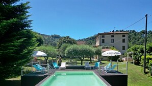 Ein Schwimmbecken mit Liegestühlen und Sonnenschirmen, umgeben von Grünpflanzen und einem Gebäude im Hintergrund.