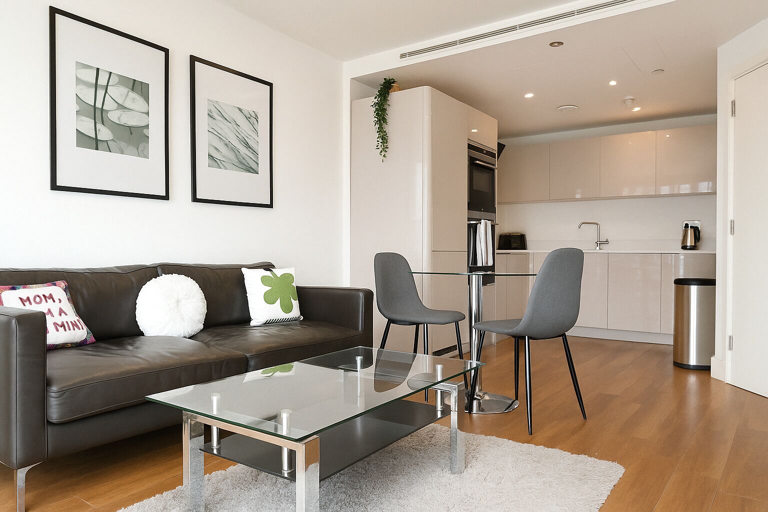 A modern living room with a glass coffee table, a dark leather sofa with decorative pillows, and a kitchen in the background.