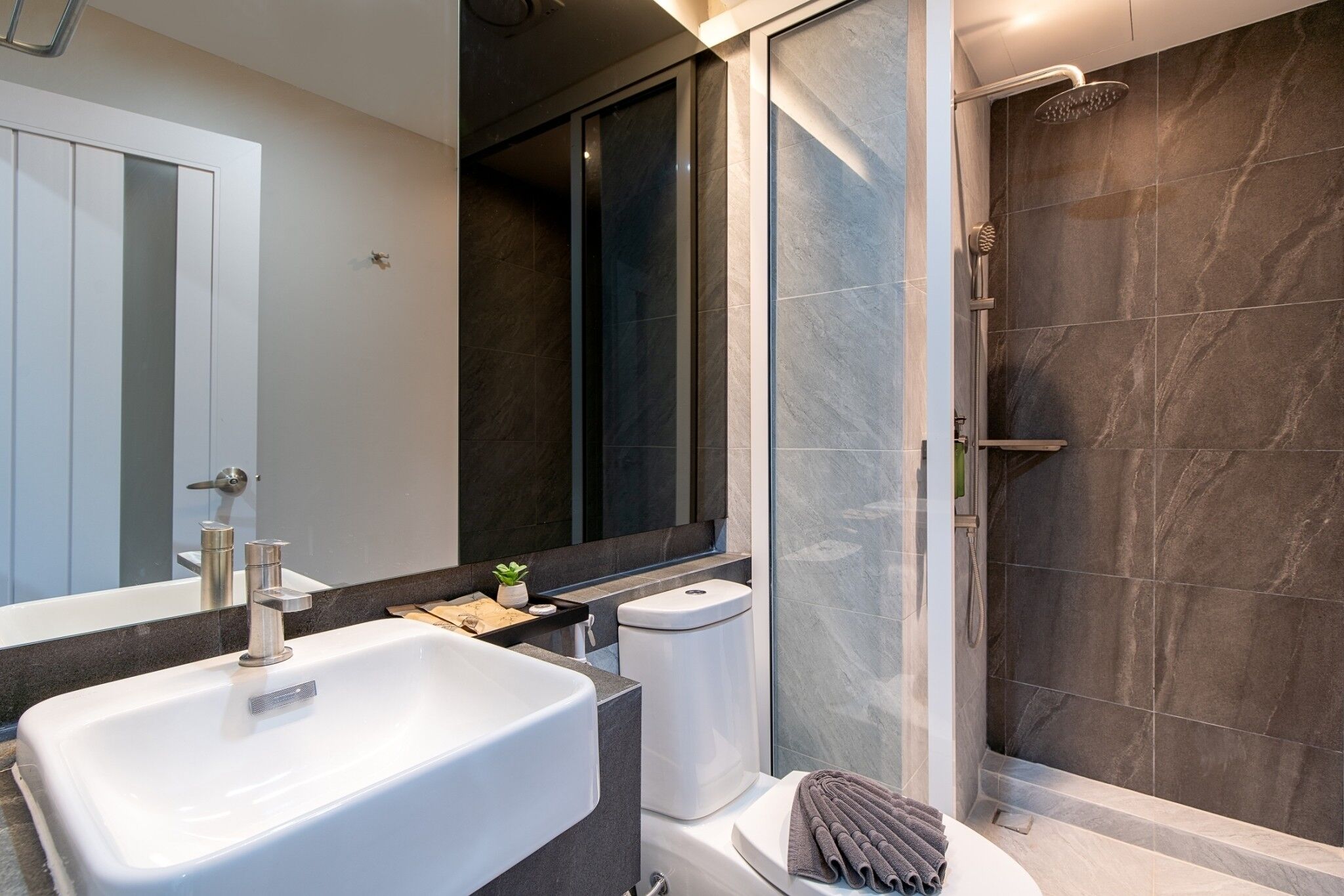 A modern bathroom with a large white sink, a glass shower enclosure, and dark marble walls.