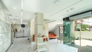 A modern hotel lobby with a marble floor, a reception desk, a seating area with a sofa and armchairs, and a glass wall offering a view of the outside. - The Riski Hotel at Bangphlat Station (Bangkok)