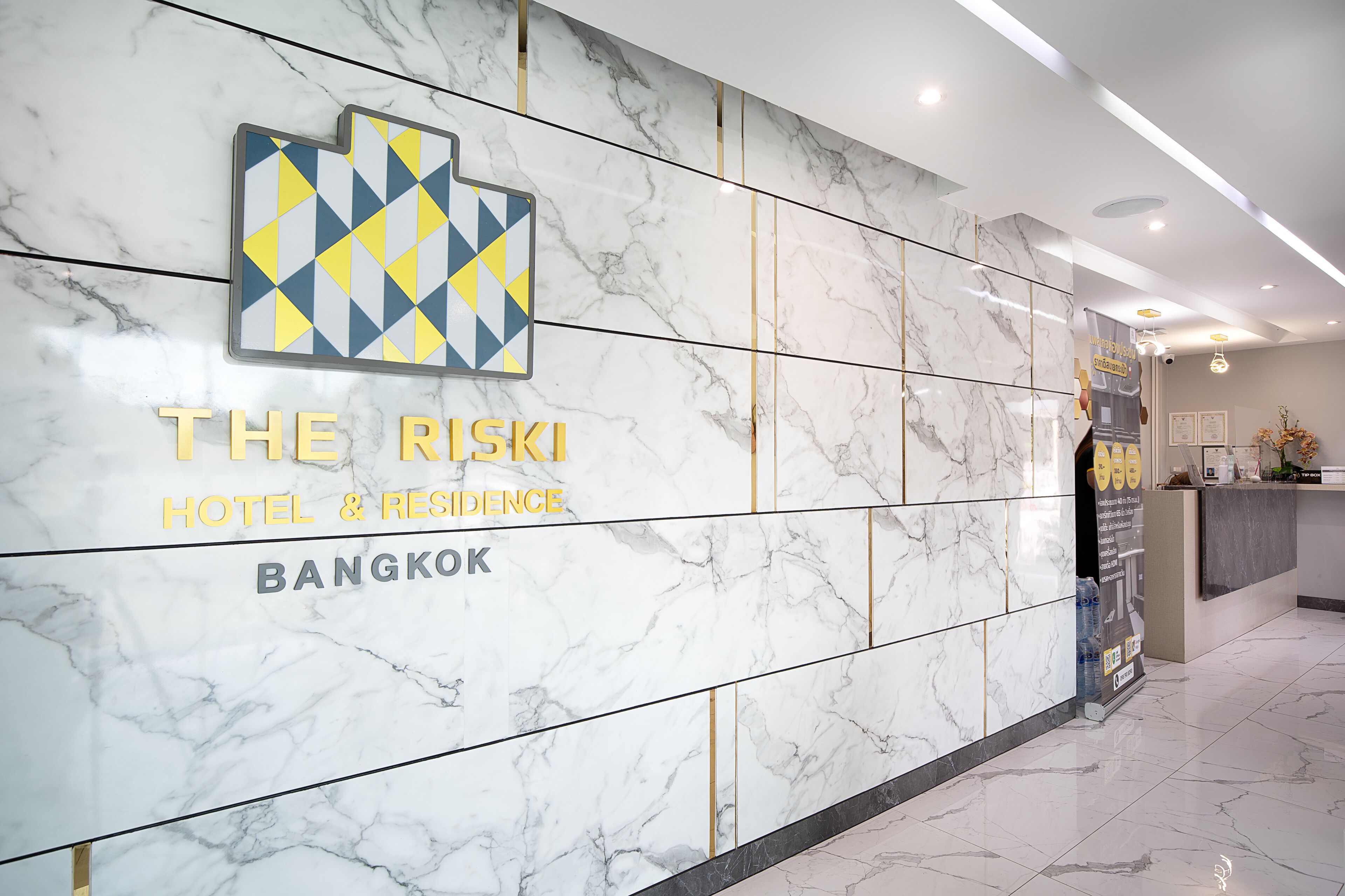 Uma área de recepção com parede de mármore, um logotipo geométrico e o texto "THE RISKI HOTEL & RESIDENCE BANGKOK".