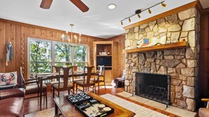 A cozy living room with a stone fireplace, wooden furniture, and a large window offering a view of trees.