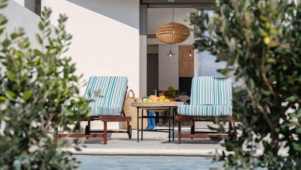 An outdoor seating area with two striped chairs and a table, a wicker hanging light, and a view of the building's interior through a glass door. - Naxos Blue Dream Villa With Pool (Naxos)