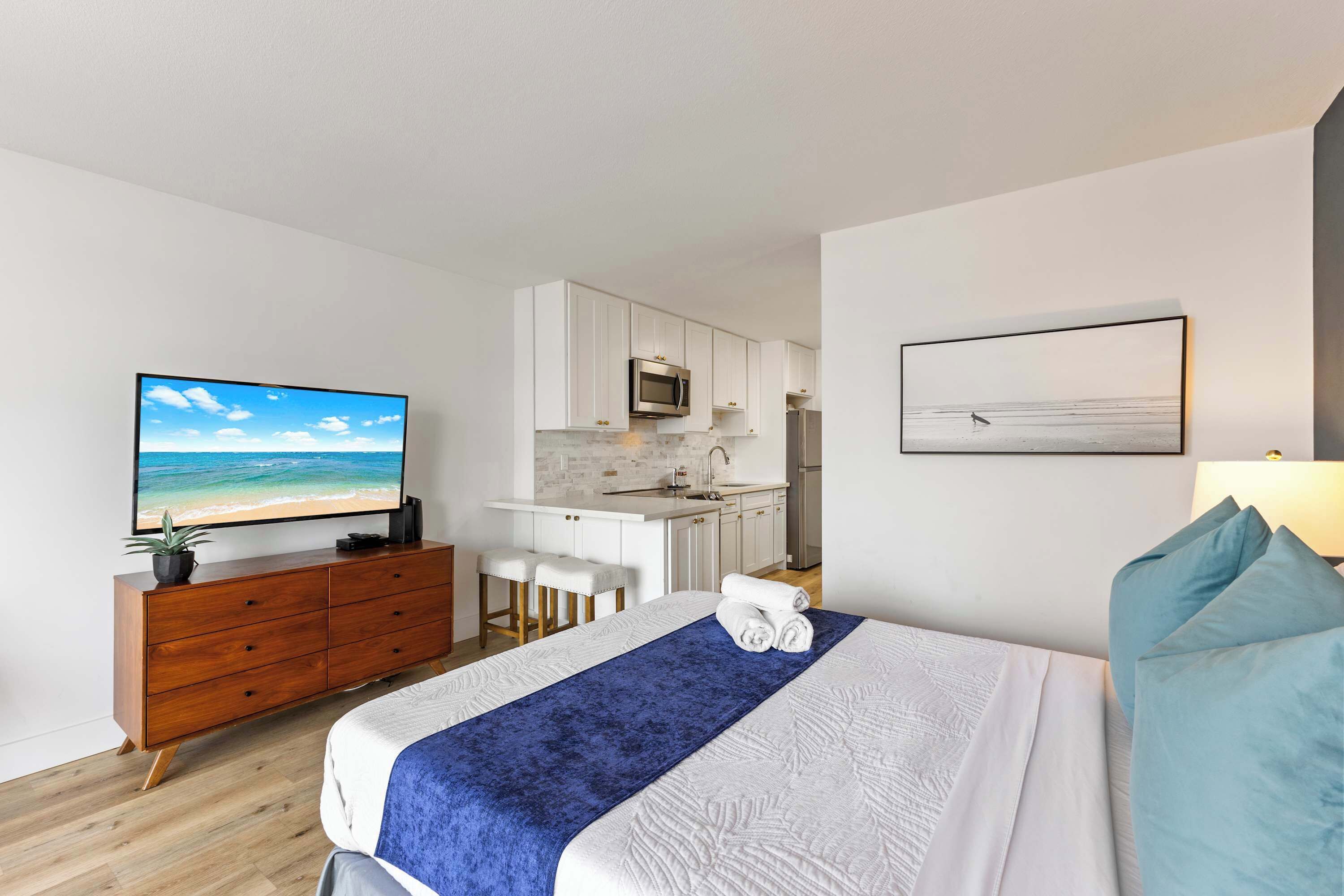 A modern hotel room with a bed, a TV, a kitchenette, and a view of the ocean.