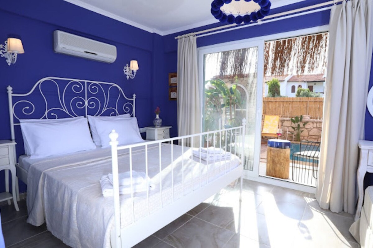 A bedroom with a bed, a nightstand, a window with curtains, and a view of an outdoor swimming pool.