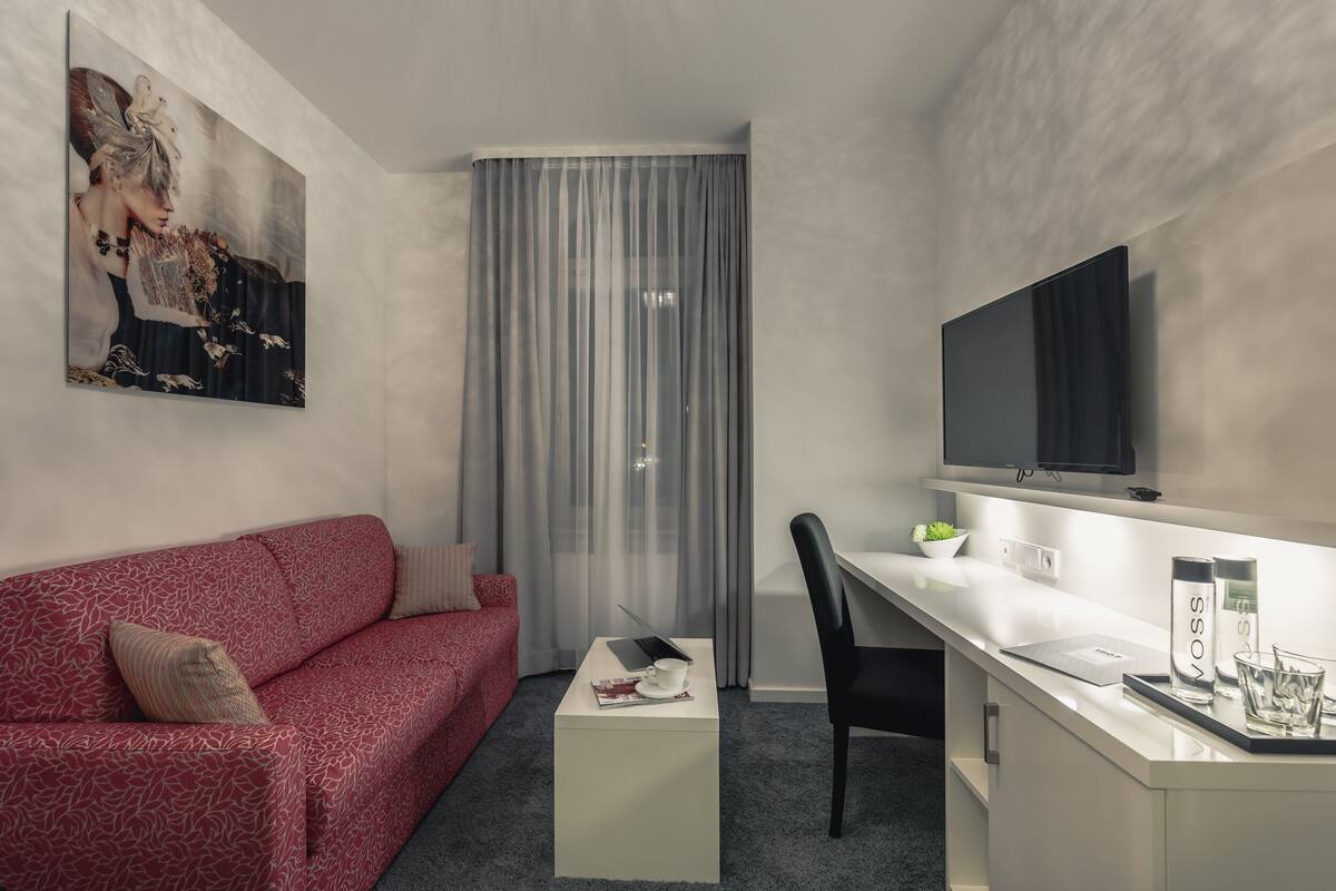 Una habitación de hotel moderna con un sofá rojo estampado, un escritorio blanco con una computadora portátil, un televisor de pantalla plana y un ventanal con cortinas translúcidas.