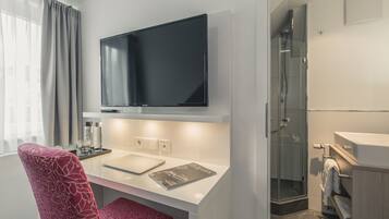 Habitación de hotel moderna con una televisión de pantalla plana sobre un escritorio blanco, zona de ducha y lavamanos blanco.