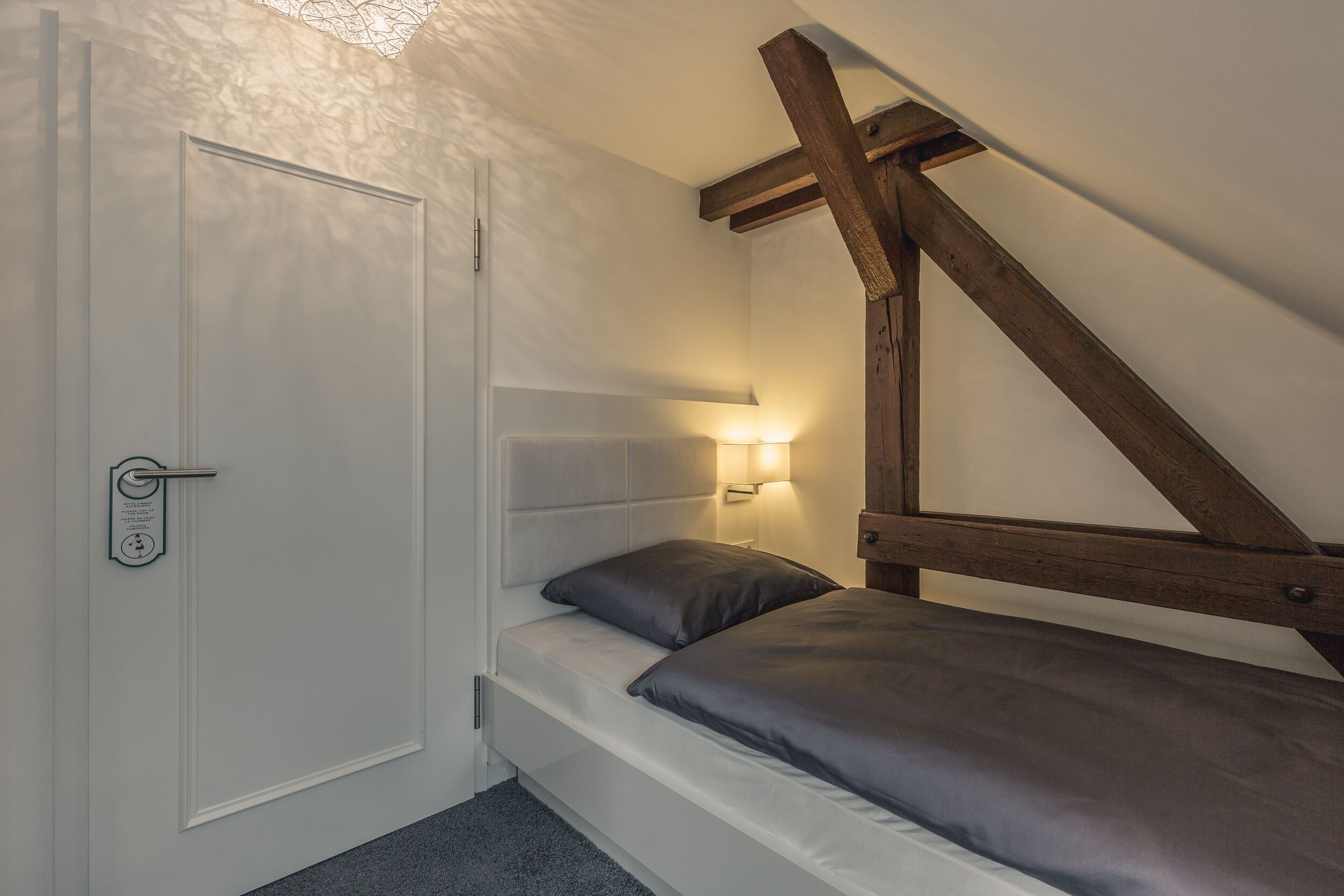 Een kleine kamer met een enkel bed, een schuine kap, een houten balkenstructuur, een witte deur met slot en een wandlamp.