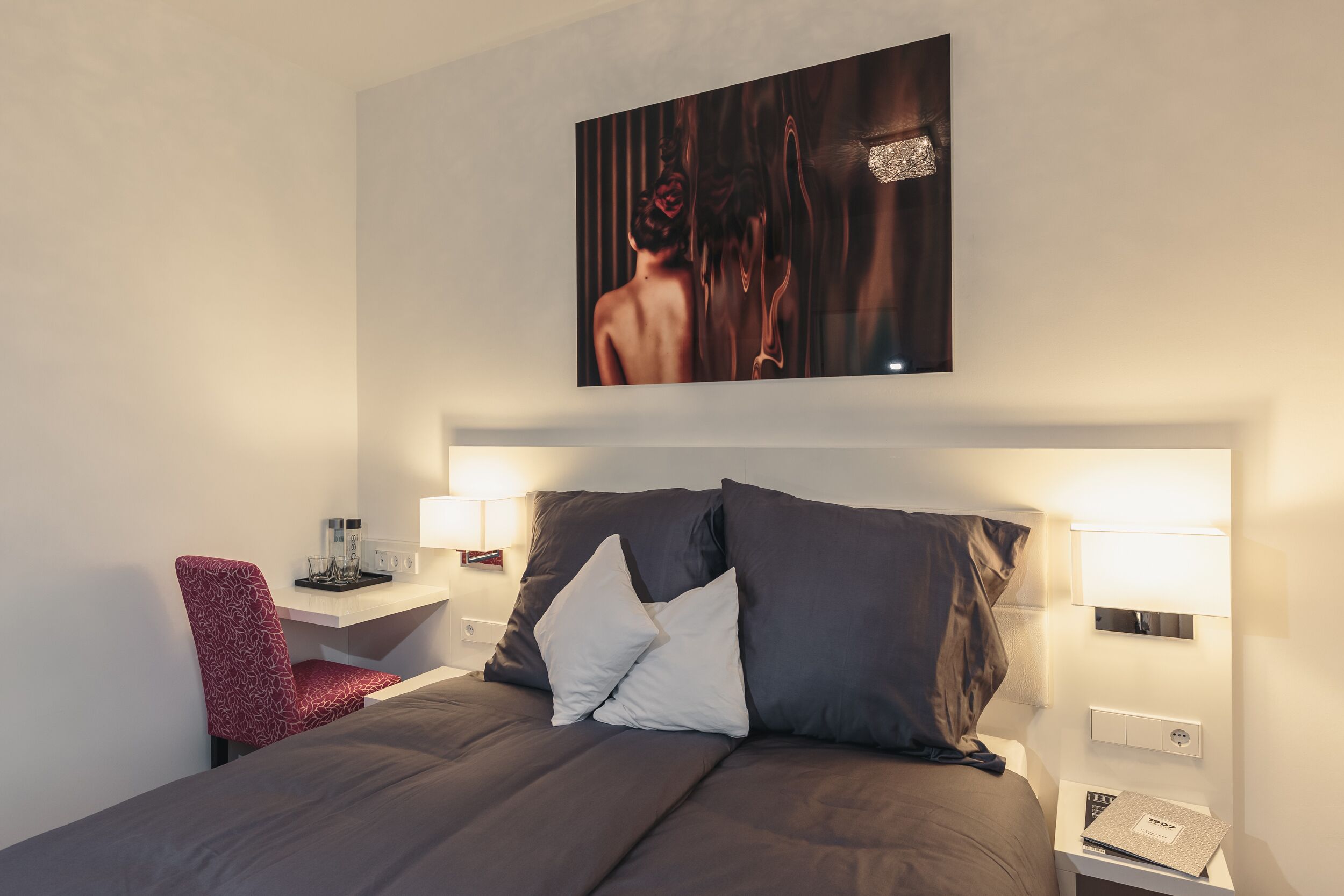 A modern hotel room with a large bed, two bedside tables, a red chair, and a wall-mounted artwork.