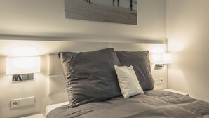 Un lit bien fait, avec une tête de lit, deux lampes de chevet et un tableau accroché au mur représentant des personnes sur une plage.