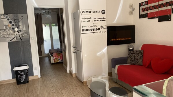 A modern living room with a red sofa, a glass coffee table, and a flat-screen TV mounted on the wall. There is a small table with a candle and a decorative item on the left. A door leads to a balcony with a view of the outside. - Apartment T2 classified 3* terrace 150 m from the beach Toulon Mourillon (Toulon)
