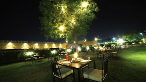 Un espacio para comer al aire libre con mesas y sillas, iluminado con luz ambiental, frente a un edificio histórico y un gran árbol.