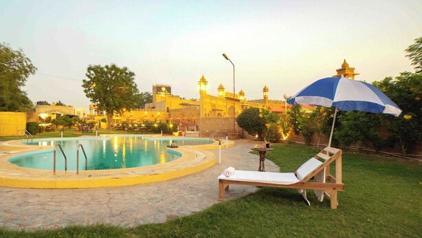 Una piscina con una tumbona y una sombrilla, rodeada por un jardín ajardinado y un edificio con una cúpula y minaretes al fondo.