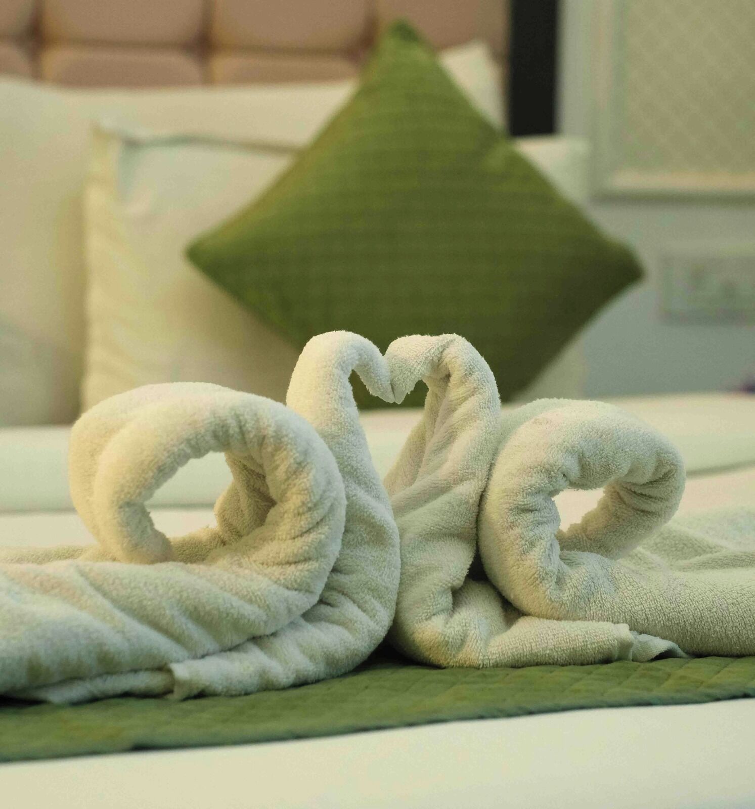 A bed with a green pillow and two white towels arranged in a heart shape.