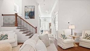 A spacious living room with a wooden staircase, white sofas, and a large painting on the wall. - LSV/Golf Cart, Private Pool, Fire Pit! Steps to Beach! Central Seagrove! Gulf Views!! (Santa Rosa Beach)