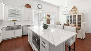 A modern kitchen with white cabinetry, a marble island, and a dining area with white chairs. - LSV/Golf Cart, Private Pool, Fire Pit! Steps to Beach! Central Seagrove! Gulf Views!! (Santa Rosa Beach)