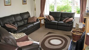 A living room with two black leather sofas, a floor lamp, and a large window with curtains. - Popular Comfortable Well Equipped Cottage in Peaceful Ullapool Street w Parking (Ullapool)