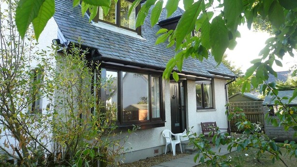A two-story house with a sloped roof, large windows, and a small porch with a chair. - Popular Comfortable Well Equipped Cottage in Peaceful Ullapool Street w Parking (Ullapool)