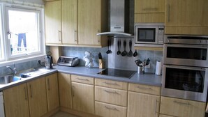 A kitchen with wooden cabinets, a microwave, an oven, a toaster, and various utensils on the counter. - Popular Comfortable Well Equipped Cottage in Peaceful Ullapool Street w Parking (Ullapool)