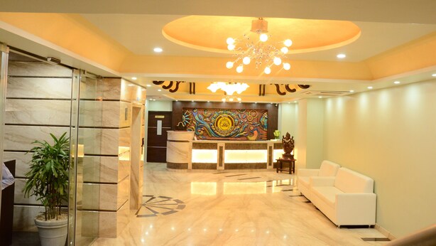 A hotel lobby with a reception desk, a white sofa, a decorative wall, and a chandelier. - JAI MAATA GRANDEUR (Shimoga)