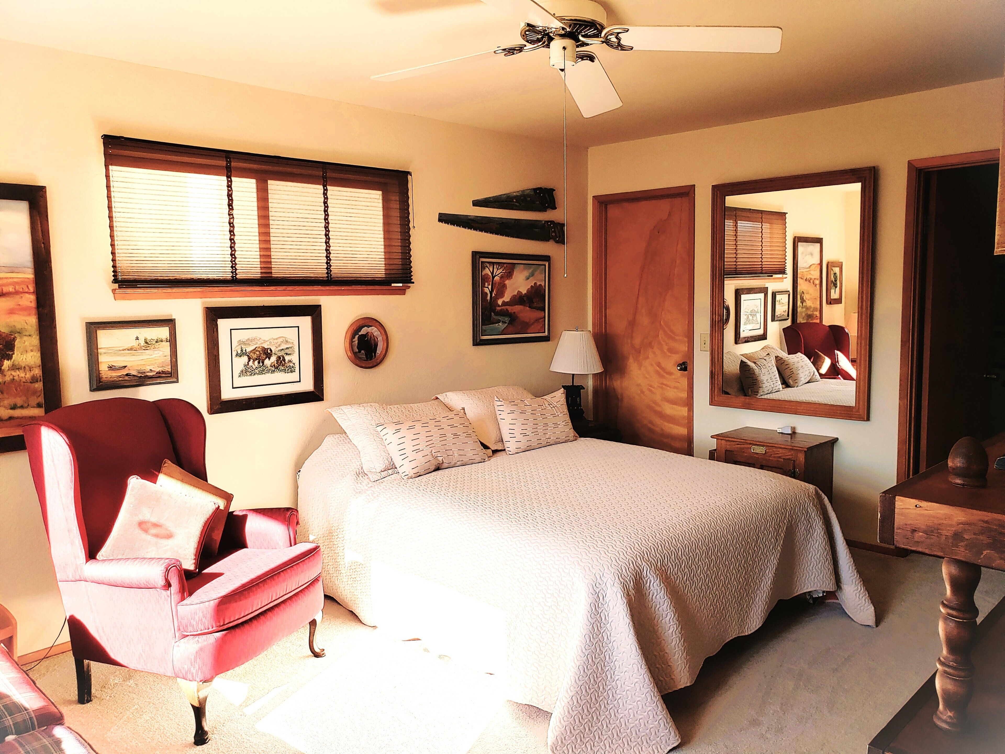Quarto com cama, poltrona vermelha, mesa de cabeceira de madeira, ventilador de teto e quadros na parede.