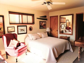 Une chambre à coucher avec un lit, un fauteuil rouge, une table de chevet en bois, un ventilateur de plafond et des tableaux encadrés accrochés au mur.