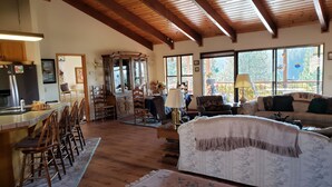 Amplia sala de estar con techo de madera, comedor con sillas de madera, sofá y un ventanal con vistas a árboles.