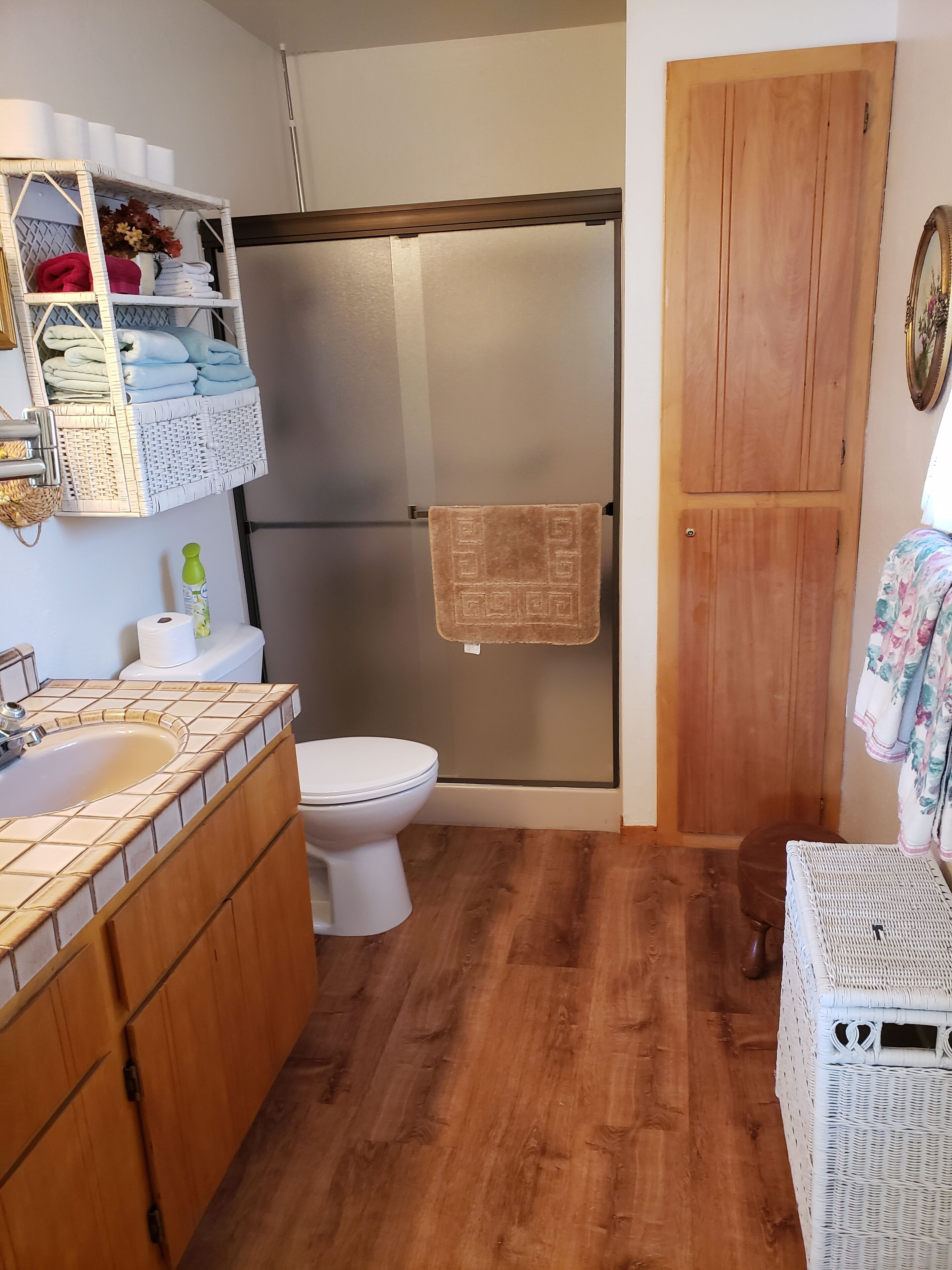 A bathroom with a wooden floor, a white toilet, a sink with a tiled countertop, a shower with a glass door, and a wooden door.