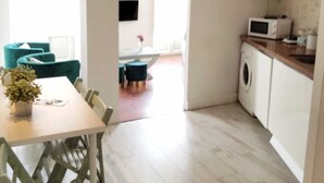 Una cocina moderna con microondas, lavadora y televisor montado en la pared. Hay una zona de comedor con mesa y sillas, y una sala de estar con sofá y planta.