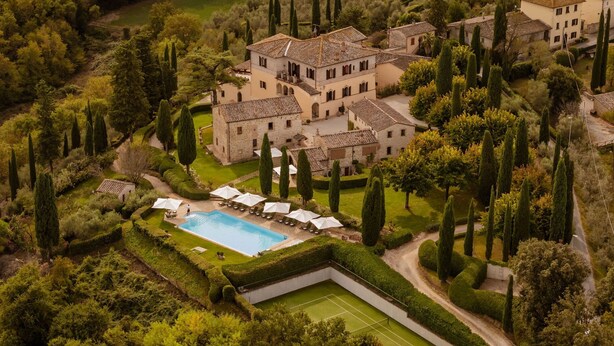 An aerial view of a large estate with a swimming pool, tennis court, and multiple buildings surrounded by lush greenery and tall cypress trees.