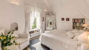 A bedroom with a large bed, a bedside table, a chair, a window with curtains, a mirror, a bookshelf, and a statue.