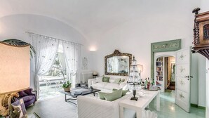 A spacious living room with a sofa set, a coffee table, and a chandelier. There is a large mirror on the wall and a bookshelf filled with books.