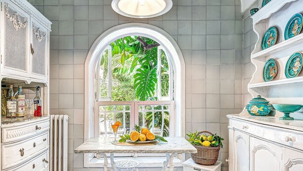 A kitchen with a white table, a window with a view of greenery, and a shelf with decorative plates.