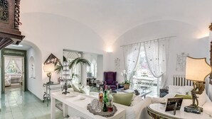 A spacious living room with a white sofa, a coffee table, and a dining table. There is a purple armchair and a chandelier. The room has large windows with white curtains.